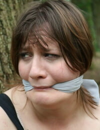 Fat female is gagged and tied to a tree in the woods with her clothes on